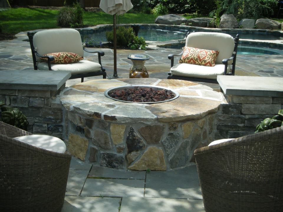 round brick firepit with several chairs around it and pool in background