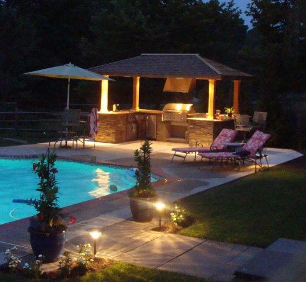 covered outdoor kitchen lit up at night by a pool. Beach chairs and umbrella next to it